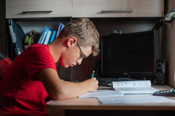 A teenage boy in a red T-shirt sits at a table at home and does his homework. A boy with glasses writes in a notebook and looks at a textbook. Study without gadgets. - Foto, Bild