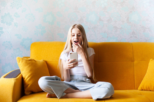 Aufgeregtes Mädchen blickt in ihr Smartphone und liest auf einem gelben Sofa eine Nachricht. Verkaufsförderung, gute Nachrichten, Online-Gewinn, aktuelles Konzept. Selektiver Fokus. - Foto, Bild