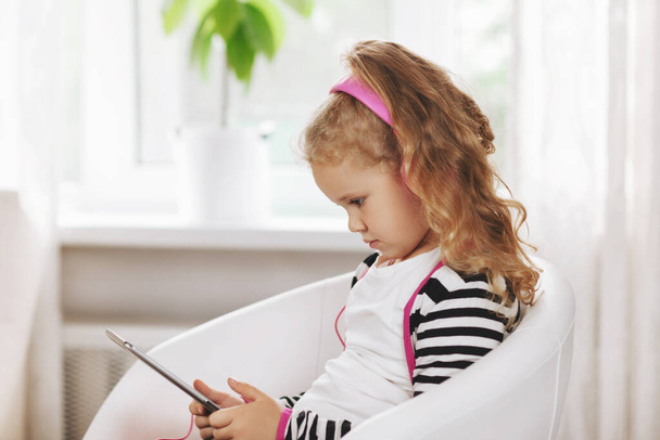 Egy rózsaszín fülhallgatós iskolás lány ül egy táblagéppel, és egy online leckét hallgat. Online oktatás, online képzés, közösségi távolság - Fotó, kép