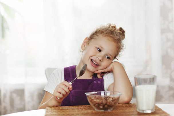 Cute little baby girl having Breakfast cereal with milk in the kitchen. The girl holds a spoon in her hand, there are cereals and a glass of milk on the table. Healthy Breakfast - Photo, Image