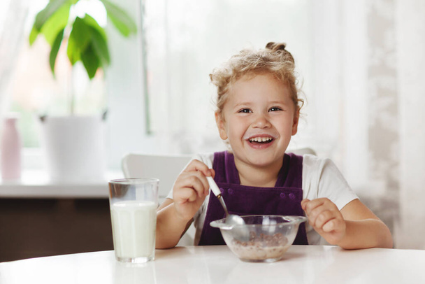 Charmante Mädchen isst Müsli mit Milch zum Frühstück und. Auf dem Tisch liegen Schokoladenflocken, ein Glas Milch. Gesundes Frühstück, Betreuung der Kinder. Raum für Text - Foto, Bild