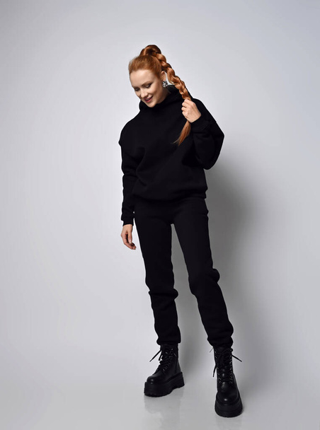 Smiling woman in trendy sports suit hoodie, pants and brutal shoes stands holding her braid with hand, looking down - Zdjęcie, obraz