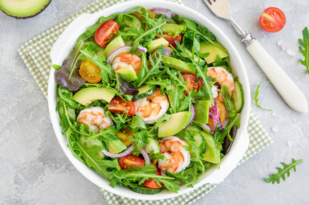 Delicious fresh salad with prawns, tomatoes, avocado, cucumber and onions in a bowl on a gray concrete background. Healthy dish. Top view, copy space - Photo, Image