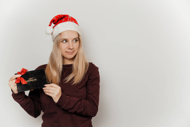 Bordo kazaklı ve kırmızı Noel Baba şapkalı bir kadın gri arka planda siyah hediye sertifikası taşıyor. - Fotoğraf, Görsel