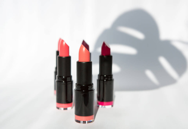 colored bright lipstick of various pink shades on a white background with shadows of leaves and bright lipstick. background for makeup artist. articles and advertising of cosmetics. copy space. - Photo, Image