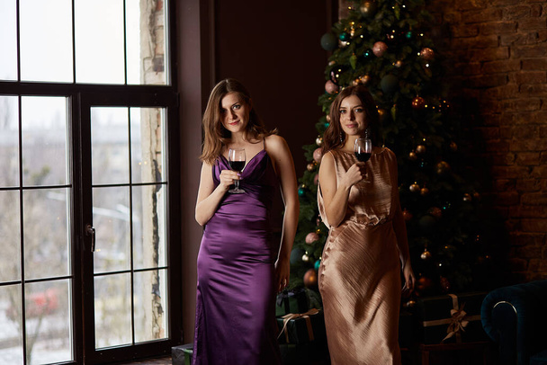 Onnellisia valkoihoisia naisia viinilasillisten kanssa joulukuusen lähellä. Stylishevening mekot, kampaus ja muodostavat. Juhlava kodin sisustus. Uudenvuoden juhlakonsepti. - Valokuva, kuva