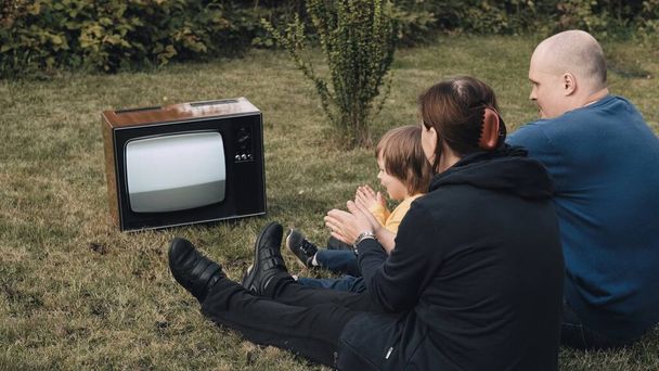 La familia está sentada en la hierba y viendo la vieja televisión retro. Aplauden. - Foto, Imagen