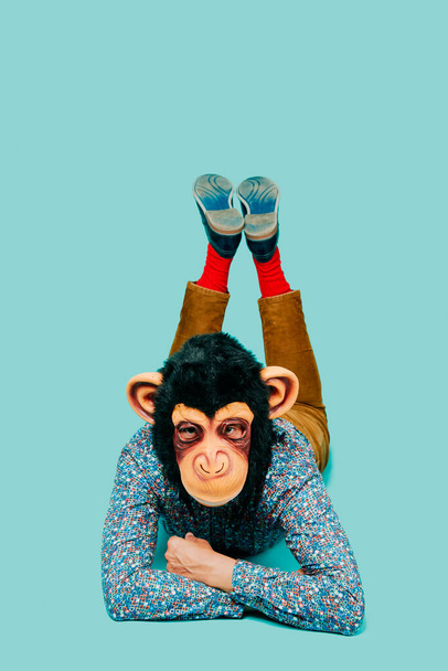 een jonge man, met een apenmasker op, kijkt vooruit terwijl hij met zijn gezicht op een blauwe achtergrond ligt met wat lege ruimte bovenop - Foto, afbeelding
