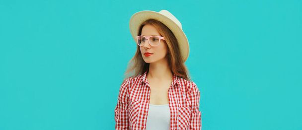 Portrait of modern young woman wearing a straw summer hat, checkered shirt looking away on blue background - Фото, изображение
