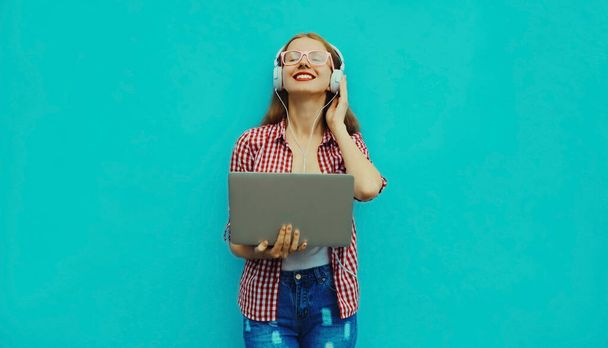 Portrait of modern young woman working with laptop listening to music in headphones on blue background - Фото, изображение