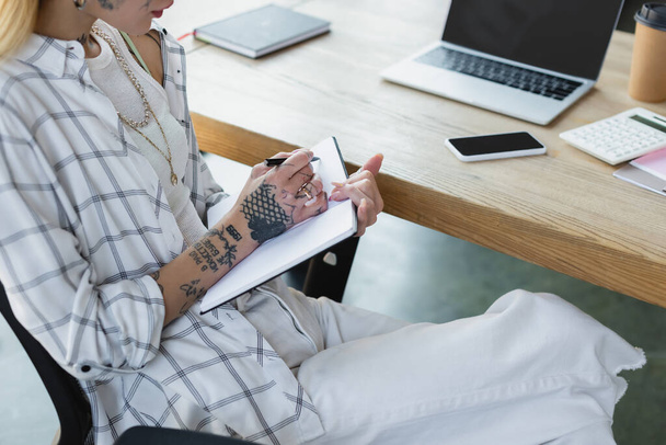 vista ritagliata della donna d'affari tatuata che scrive nel taccuino vicino ai gadget con schermo bianco sulla scrivania - Foto, immagini