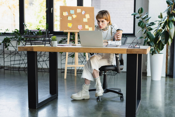 stylish tattooed businesswoman holding coffee to go while sitting at desk near laptop - Photo, Image