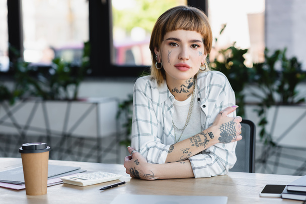 young tattooed businesswoman sitting with crossed arms at workplace in office - Photo, Image