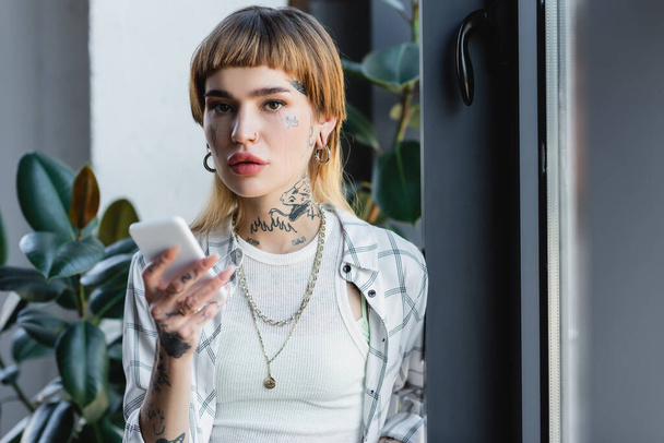 tattooed and stylish woman holding smartphone while looking at camera in office - Photo, image