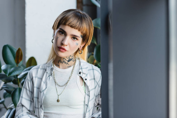 mooie jonge vrouw met tatoeage, piercing en zilveren kettingen op zoek naar camera in kantoor - Foto, afbeelding