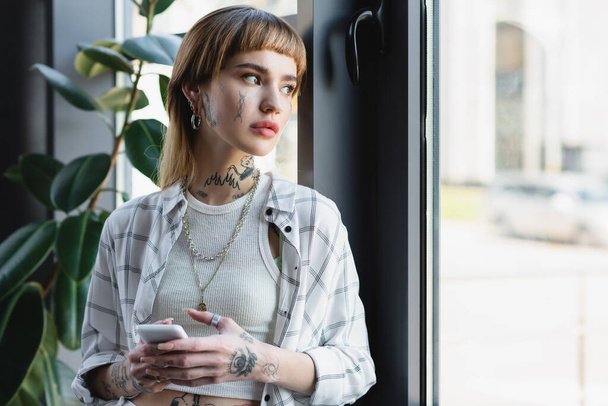 joven mujer tatuada con teléfono móvil de pie cerca de la ventana en la oficina - Foto, imagen