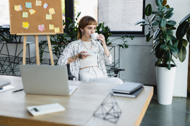 stylish tattooed woman drinking coffee while sitting at work desk near blurred laptop - Φωτογραφία, εικόνα