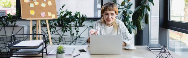 smiling businesswoman with tattoo pointing with hand during video call on computer, banner - Фото, изображение
