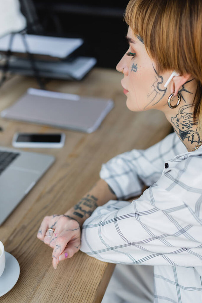 giovane donna tatuata in auricolare wireless seduta sul posto di lavoro vicino a gadget sfocati - Foto, immagini