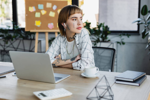tattooed businesswoman in earphone sitting at work desk near coffee cup and blurred laptop - Foto, imagen