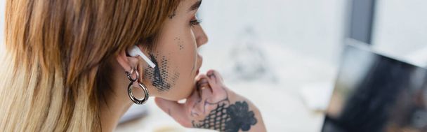young tattooed woman in wireless earphone looking at blurred laptop in office, banner - Fotó, kép