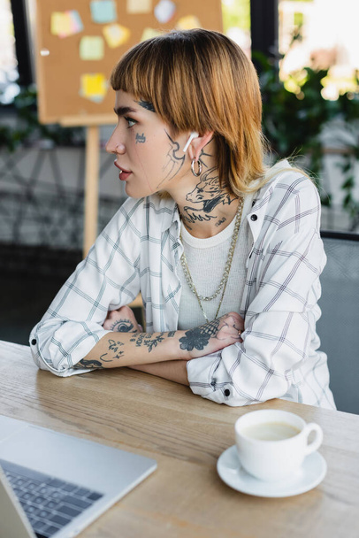 young woman with tattooed body sitting at work desk with crossed arms  - Фото, изображение