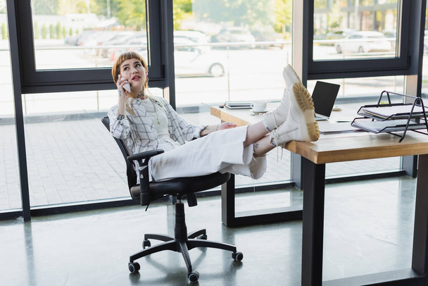 elegante donna d'affari tatuata seduta con le gambe sulla scrivania mentre parla sullo smartphone in ufficio - Foto, immagini
