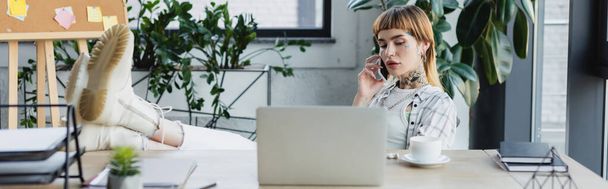 trendy businesswoman sitting with legs on work desk while talking on mobile phone, banner - Foto, Bild