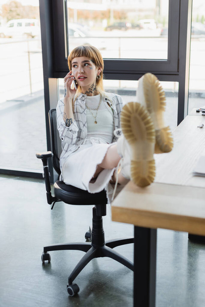 stylish tattooed woman sitting with legs on desk while talking on cellphone in office - Photo, image