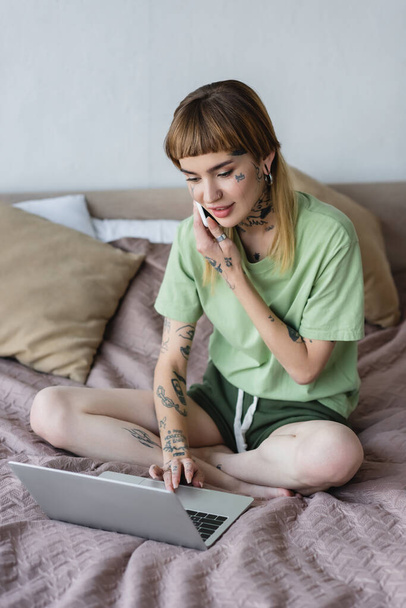 positive woman with tattooed body typing on laptop while calling on cellphone in bedroom - Fotografie, Obrázek