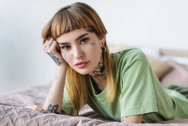 young tattooed woman looking at camera while lying on bed - Foto, Bild