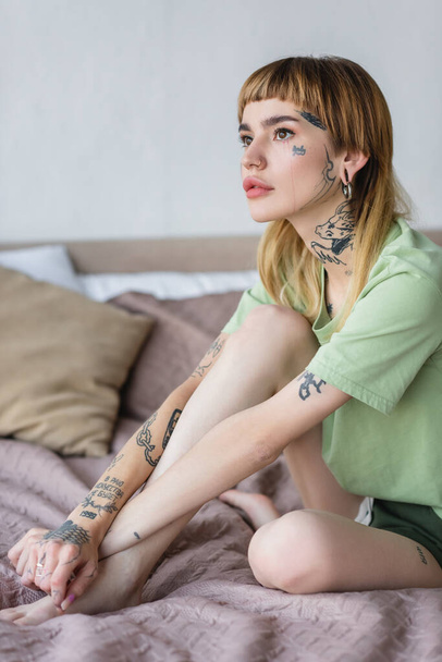 joven mujer tatuada mirando hacia otro lado mientras está sentada en la cama en casa - Foto, Imagen