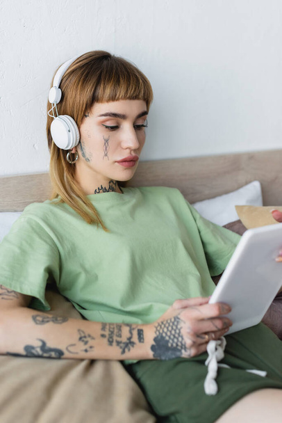tattooed woman holding digital tablet while listening online lesson in headphones at home - Фото, изображение