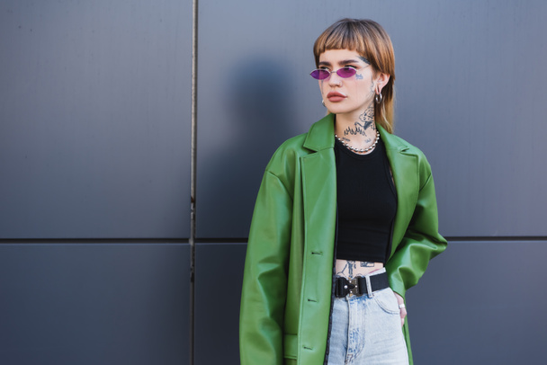 tattooed woman in green jacket standing with hand on hip and looking away near grey wall - Фото, изображение