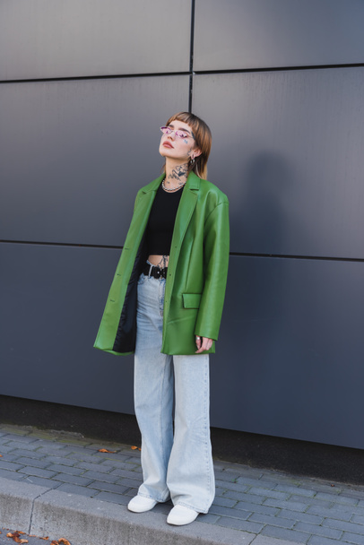 vue pleine longueur de femme tatouée en veste et jeans en cuir vert debout près du mur sur la rue de la ville - Photo, image