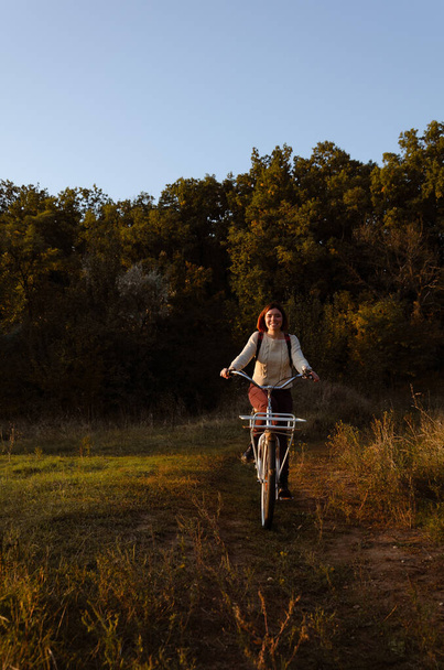 Mujer sonriente con el pelo corto en suéter blanco está montando una bicicleta en el bosque durante la hora dorada, mirando a la cámara. - Foto, imagen
