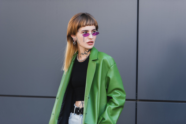 tattooed woman in green leather jacket and trendy eyeglasses looking away on city street - 写真・画像