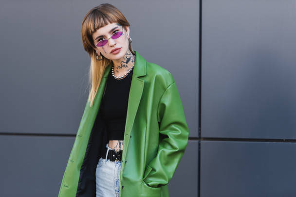 tattooed woman with piercing looking at camera while posing with hand in pocket of green jacket - Photo, Image