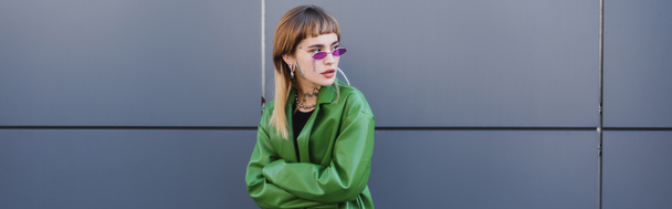 tattooed woman in stylish eyeglasses and green leather jacket looking away near grey wall, banner - Foto, afbeelding