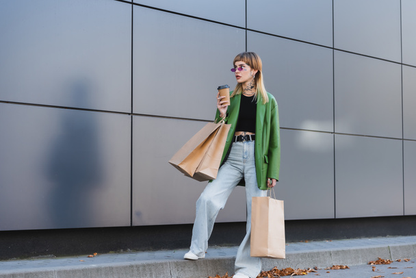 stylish tattooed woman in green jacket and jeans standing with shopping bags and paper cup outdoors - 写真・画像