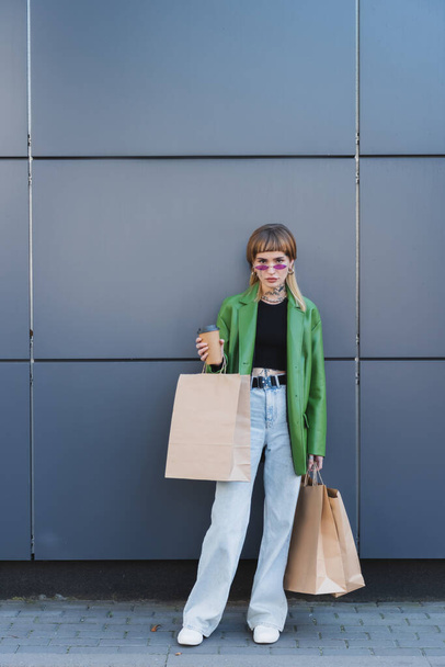 trendy tattooed woman with shopping bags and coffee to go looking at camera near grey wall - Photo, Image