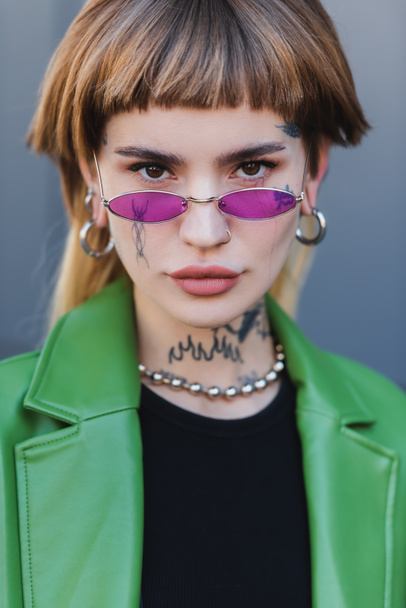 стильная татуированная женщина в очках смотрит в камеру на улице - Фото, изображение