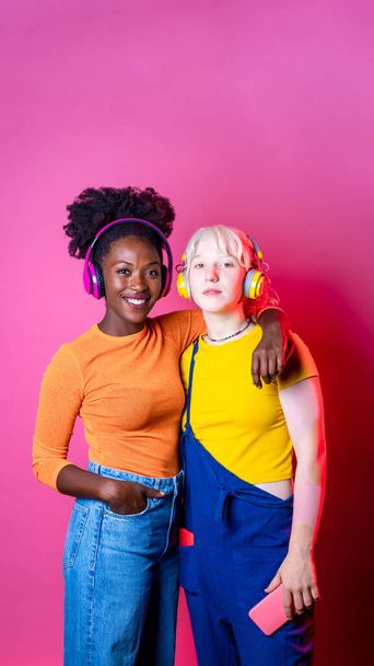 Two young diverse multicultural women best friends posing isolated background having fun laughing holding smartphone wearing wireless headphones - Foto, Bild