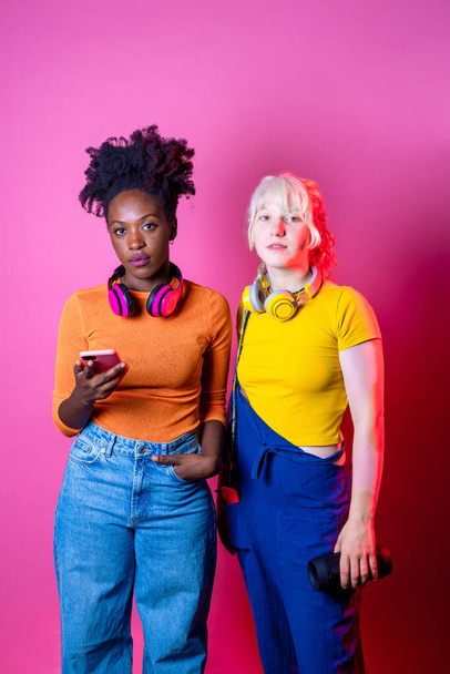 Дві молоді різноманітні мультикультурні жінки найкращі друзі позують ізольований фон, розважаючись, сміючись, тримаючи смартфон у бездротових навушниках
 - Фото, зображення