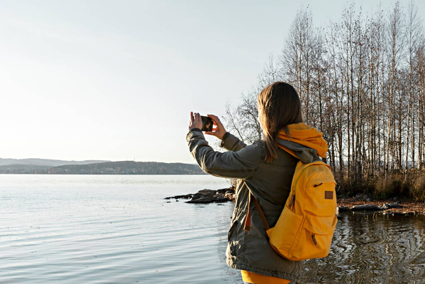 Widok z tyłu młoda blondynka z żółtym plecakiem robi zdjęcie na smartfonie jesiennego jeziora krajobrazowego, przy użyciu nowoczesnych technologii piękna w przyrodzie, jesień sezon spacery fotografowania - Zdjęcie, obraz