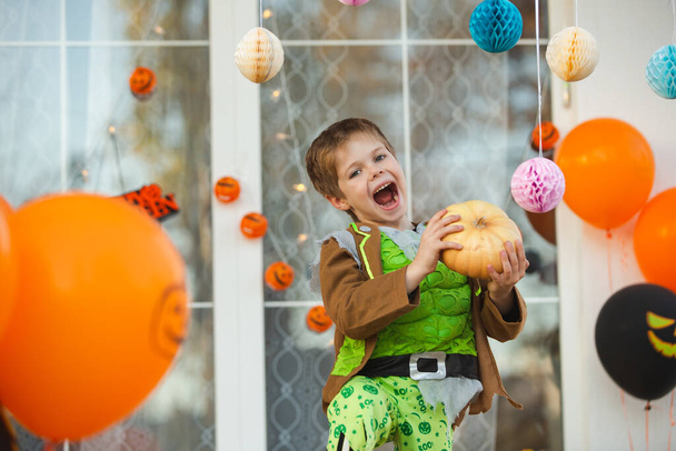 Un hermoso niño con un disfraz de zombie posando con una pequeña calabaza en sus manos. Decoraciones para la celebración de Halloween en la puerta. Acogedora terraza en una casa de campo decorada para las vacaciones - Foto, imagen
