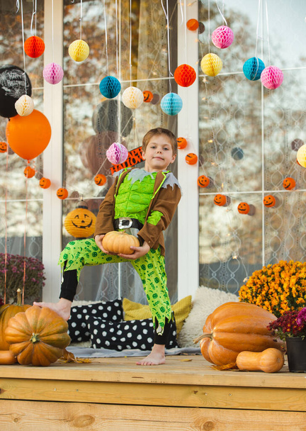 A beautiful child in a zombie costume posing with a small pumpkin in his hands. Decorations for the celebration of Halloween on the doorstep. Cozy veranda in a country house decorated for the holiday - Foto, Bild