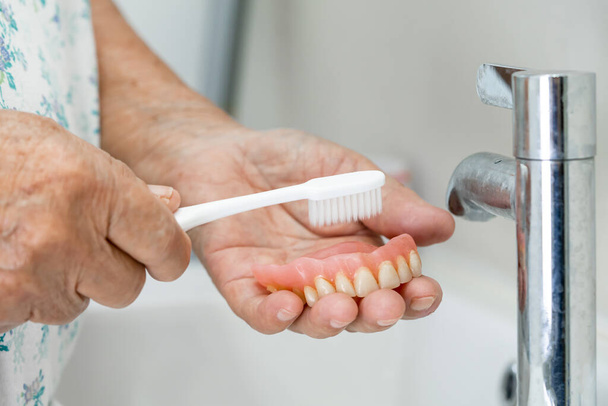 Asiática senior o anciana anciana paciente uso cepillo de dientes para limpiar parcial dentadura postiza de reemplazo de dientes. - Foto, Imagen