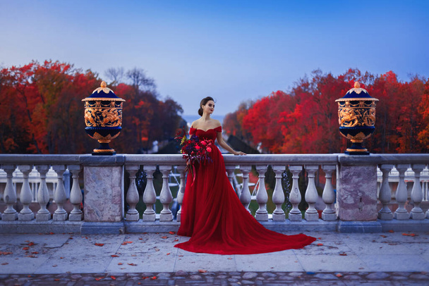 Een mooi meisje in een lange rode baljurk op het balkon tegen de achtergrond van een herfstpark in de schemering. - Foto, afbeelding