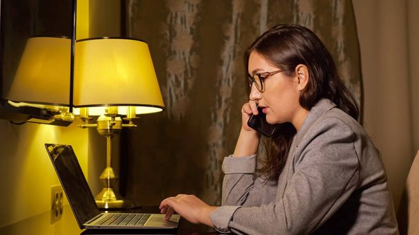 žena v obleku ve večerních hodinách pracuje na notebooku a mluví po telefonu - Fotografie, Obrázek
